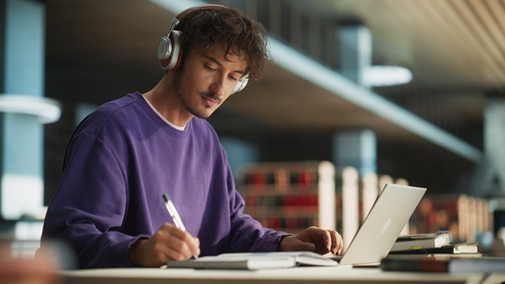 A young man wearing headphones and a purple sweatshirt is studying with a laptop and notebook in a library or study area. He is writing notes while looking at the screen, surrounded by books and soft lighting.