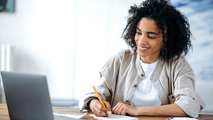 Woman at desk, writing with a pencil, whilst smiling at laptop